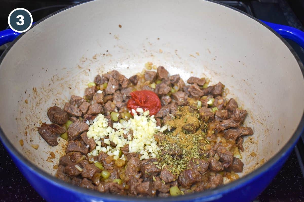 Garlic, tomato paste, and seasoning over browned steak pieces in a large pot.