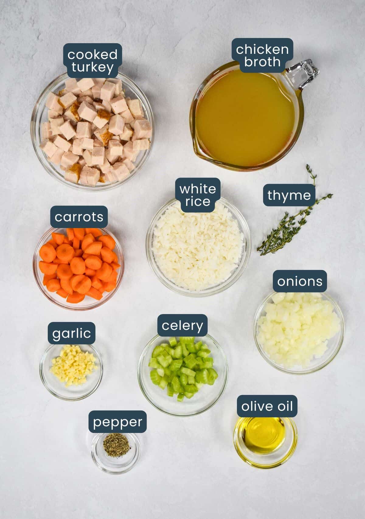 The ingredients for the soup prepped and arranged in glass bowls on a white table. Each ingredient has a label with the name above it in blue and white letters.