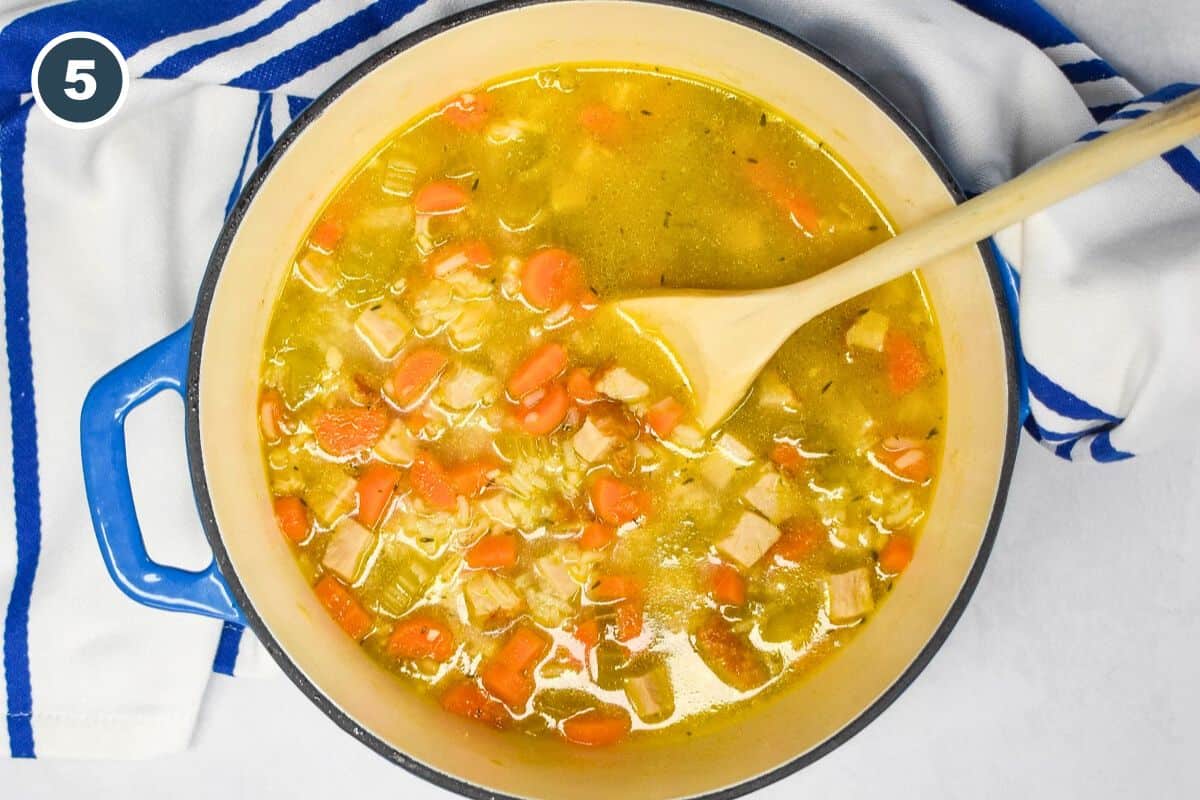 An image of the turkey and rice soup in a blue pot that is set on a white table with a blue and white kitchen towel and a wooden spoon in the pot.