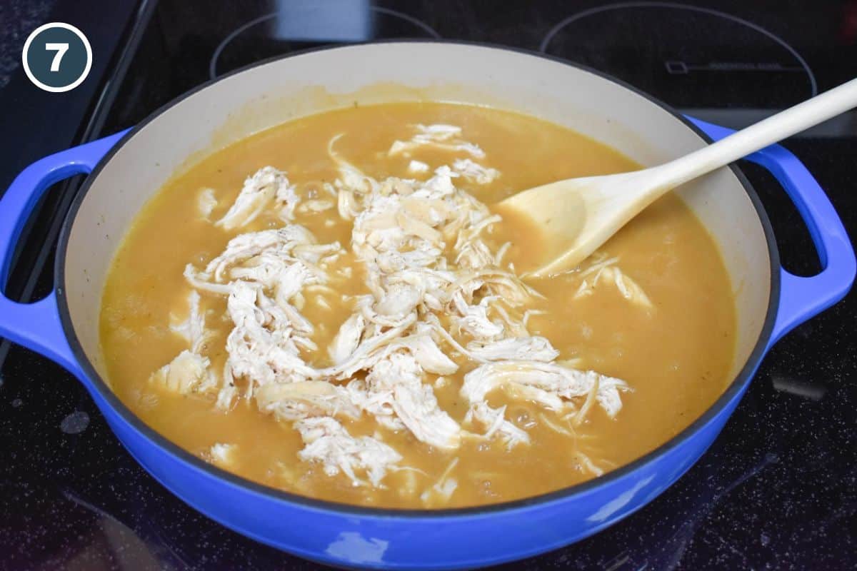 Shredded chicken added to a blue skillet containing chicken gravy, with a wooden spoon for stirring.