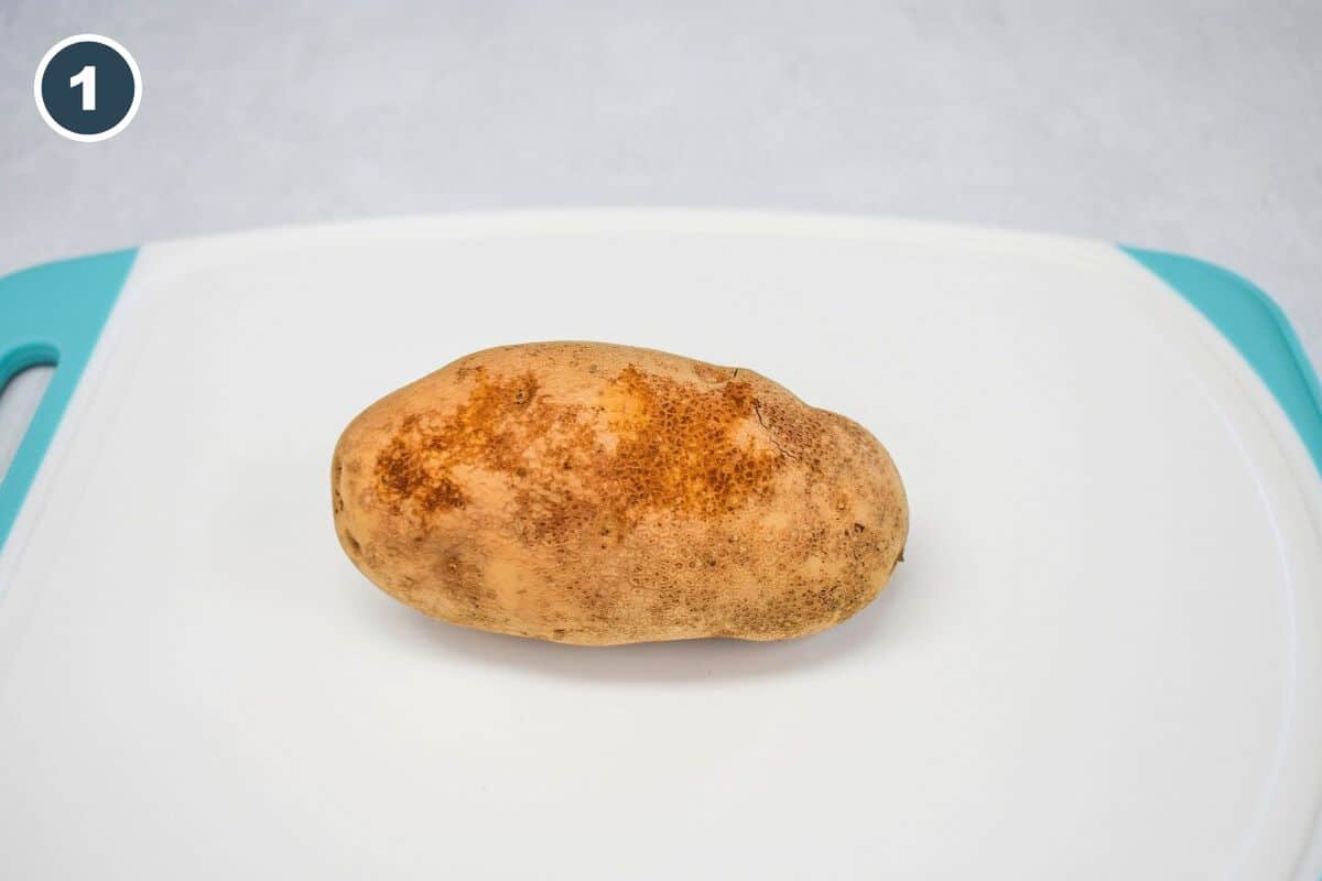 A whole russet potato sits on a white cutting board, ready to be prepped for the recipe.