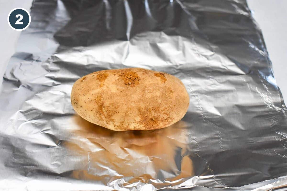 A whole russet potato sits on a sheet of aluminum foil, ready to be wrapped for baking.