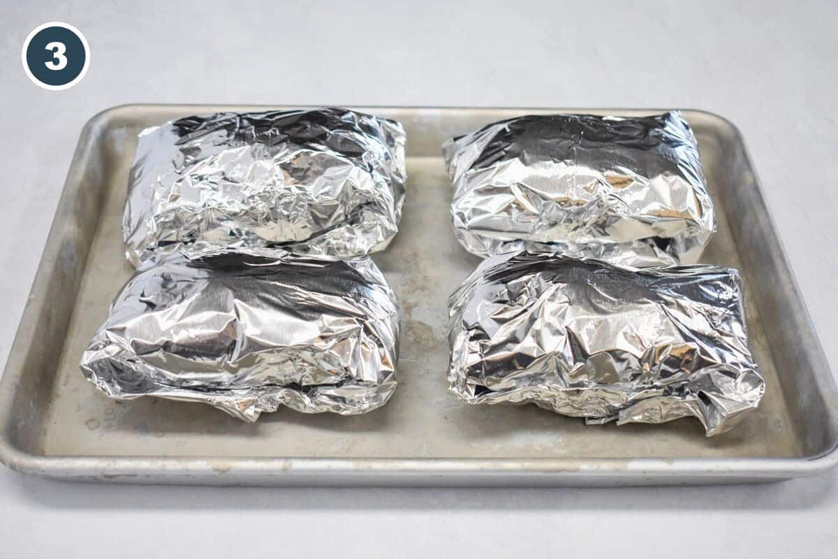 Four foil-wrapped potatoes are arranged on a baking sheet, ready to go into the oven.