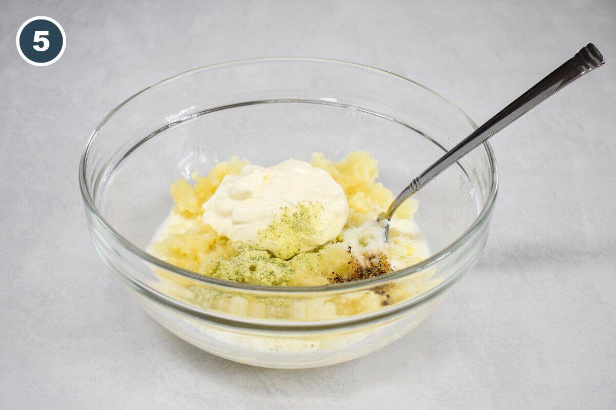 Mashed potatoes are mixed with sour cream, milk, butter, and seasonings in a glass bowl with a spoon.