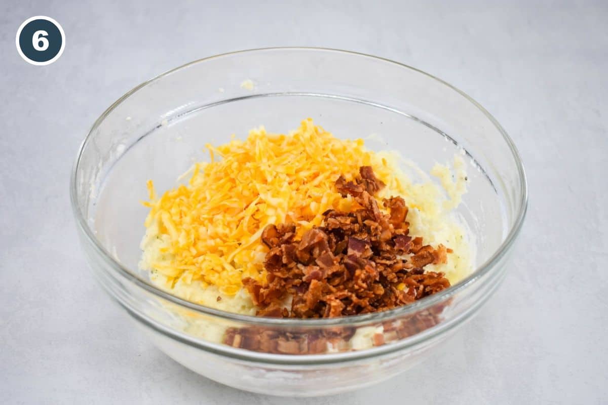 Shredded cheese and crumbled bacon are added to the mashed potato mixture in a glass bowl.