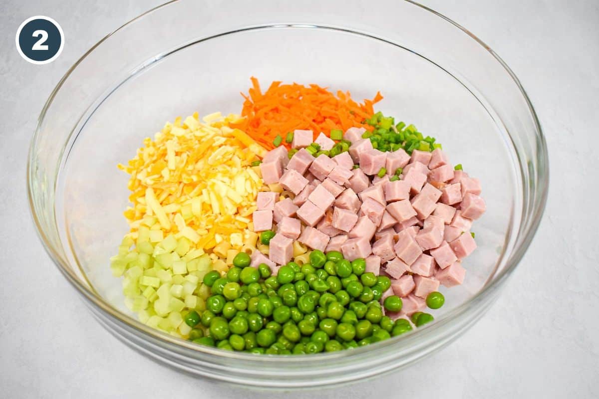 Cooked pasta covered by diced ham, shredded carrots, cheese, celery, peas, and green onions in a large, glass bowl.