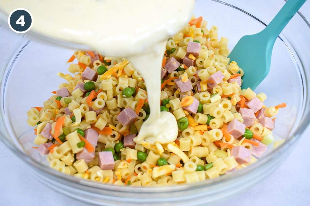 The dressing being poured over the pasta ham mixture in a large, glass bowl with a light blue spatula to the right.