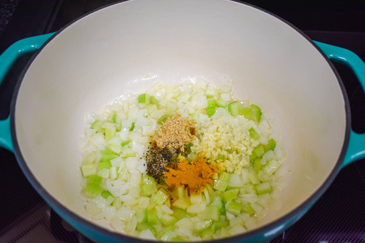 Minced garlic and spices added to diced onions and celery cooking in a large white and teal pot.