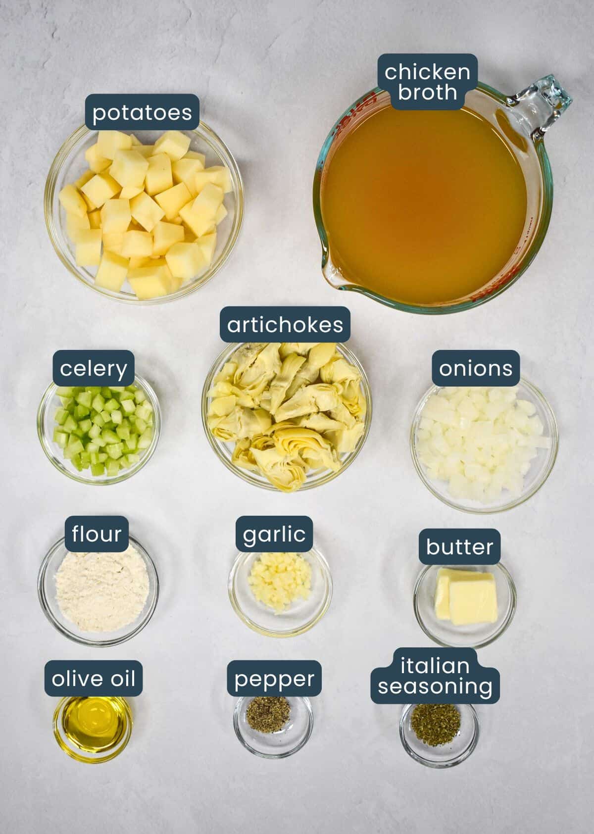 A top-down view of ingredients for artichoke soup, including diced potatoes, chicken broth, chopped artichokes, onions, celery, garlic, flour, butter, olive oil, black pepper, and Italian seasoning, all labeled in small glass bowls and measuring cups.