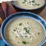 A close-up of two bowls of creamy artichoke soup, garnished with grated Parmesan cheese and fresh chopped parsley. The soup has a smooth texture and is served in rustic blue bowls on a wooden surface with the title in a text overlay.