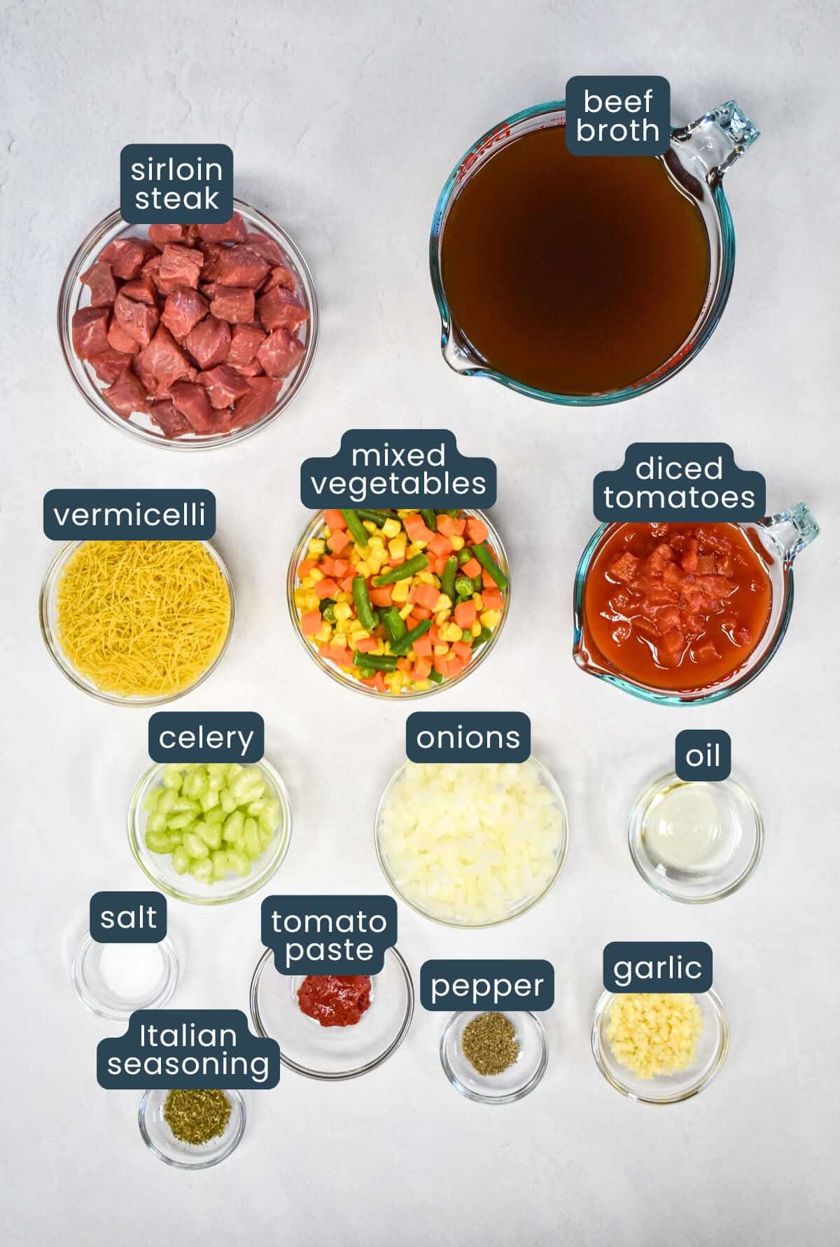 The ingredients for the soup prepped and arranged in glass bowls set on a white table with each labeled.