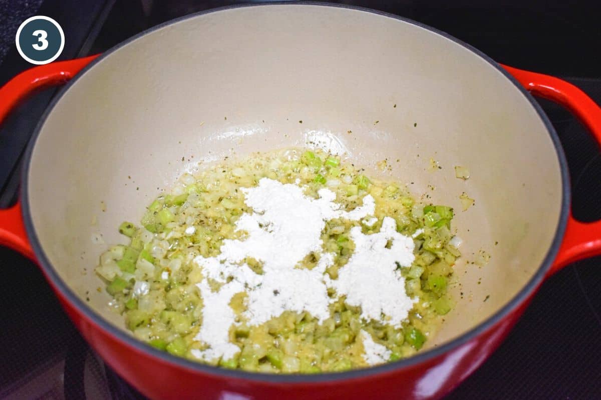 Flour sprinkled over sautéed onions, celery, and seasonings in a red enameled Dutch oven, forming a roux to thicken the artichoke soup.