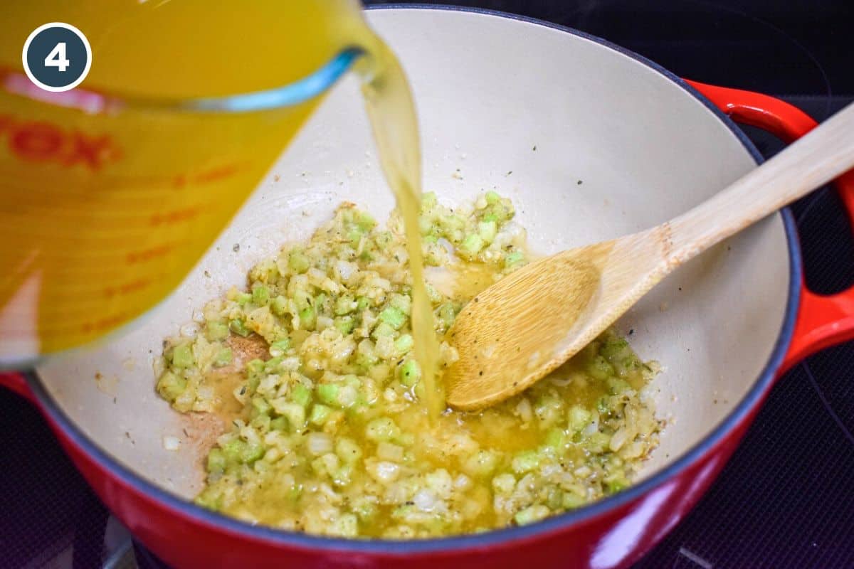 Chicken broth being poured into a red enameled Dutch oven with sautéed onions, celery, and a roux, deglazing the pot and forming the base for the artichoke soup.