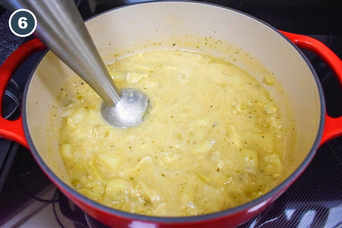 A pot of artichoke soup being blended with an immersion blender to achieve a smooth and creamy texture. The soup has thickened and taken on a golden hue.