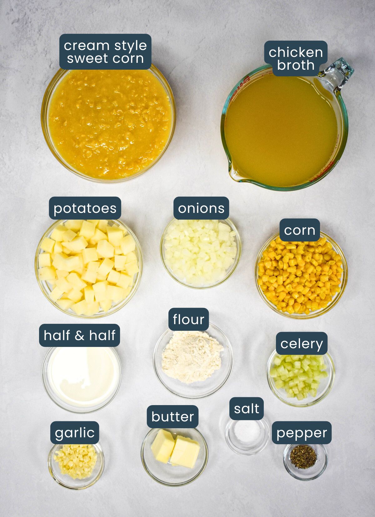 The ingredients for the corn chowder prepped and arranged in glass bowls set on a white table with each one labeled.