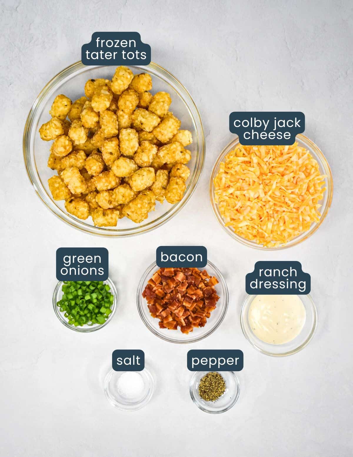 The ingredients for the loaded tater tots prepped and arranged on a white table with each labeled in blue and white letters.