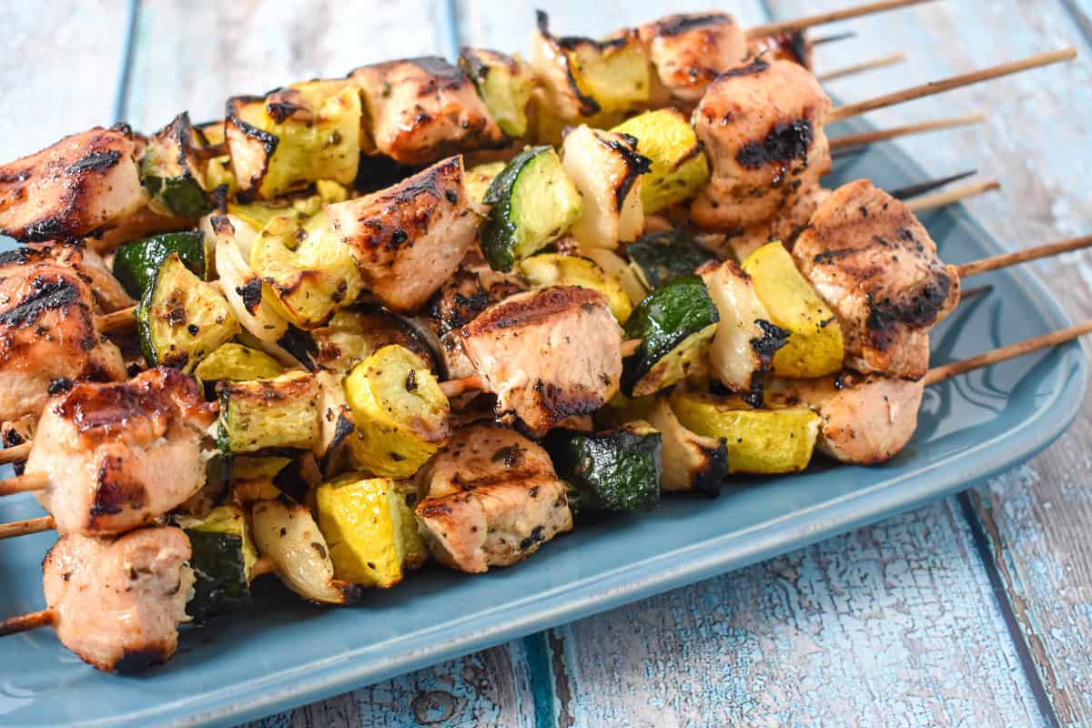 Skewers with grilled chicken, zucchini, yellow squash, and onions stacked on a blue plate.