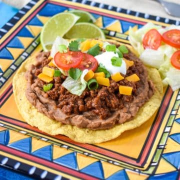 A garnished ground beef tostada served on a colorful blue and yellow plate with lime wedges and extra lettuce and tomatoes on the side.