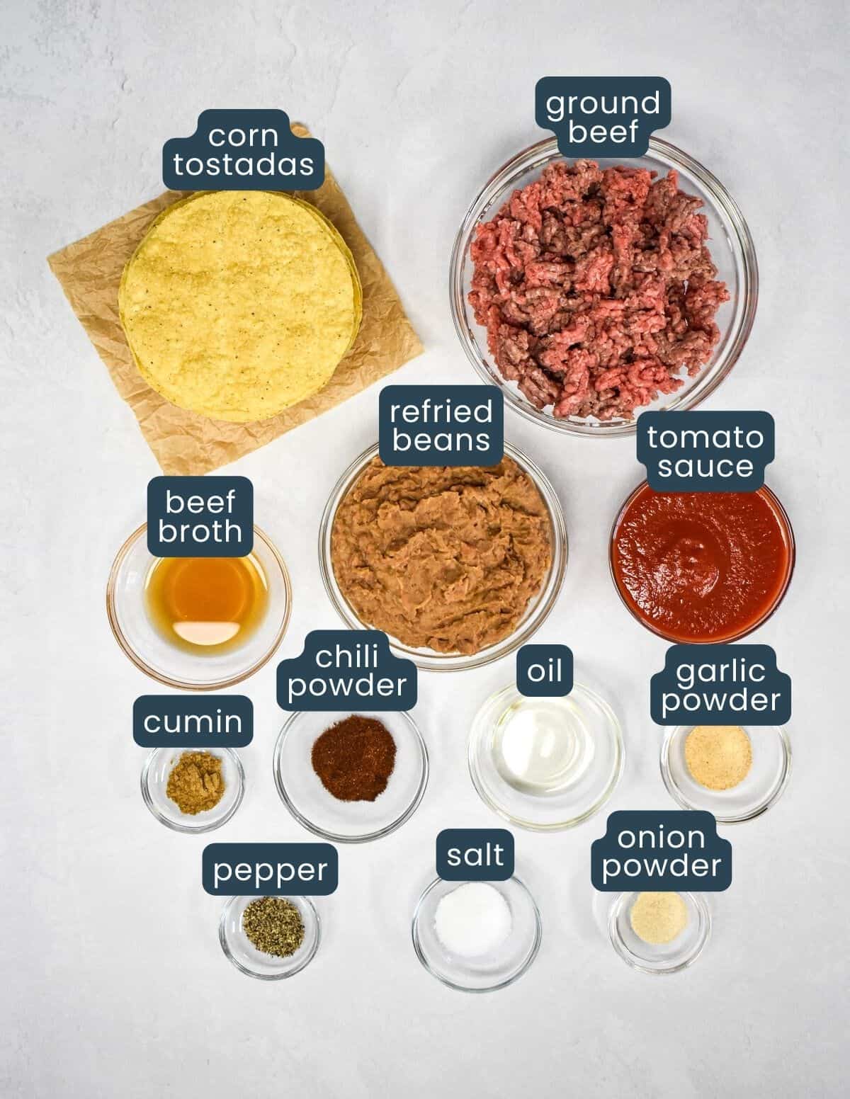 The ingredients for the tostadas, prepped and arranged in glass bowls on a white table with each labeled with blue and white letters.