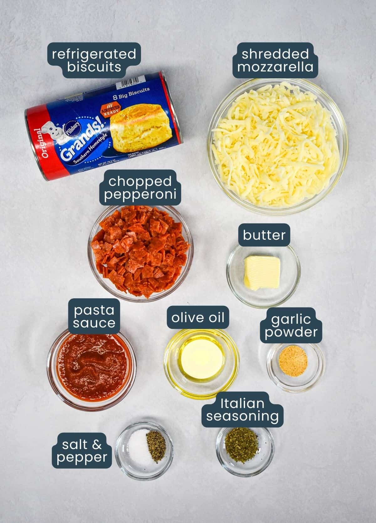 The ingredients for the pizza cups prepped and arranged on a white table with each labeled with blue and white letters.