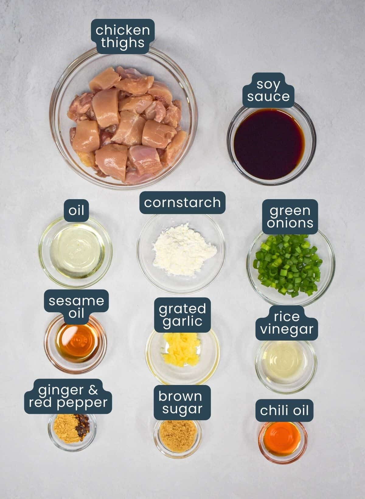 The ingredients for the sesame chicken prepped and arranged in glass bowls on a white table with each labeled with blue and white letters.