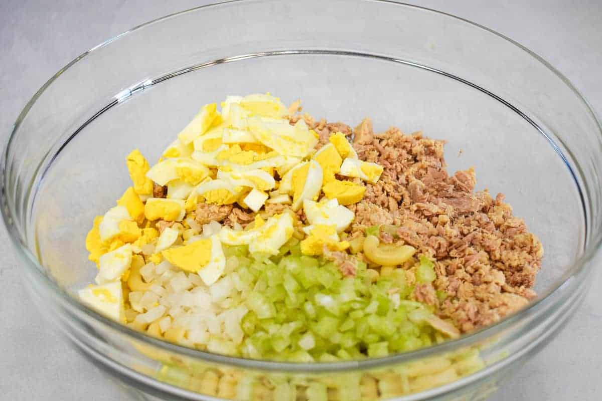 Cooked macaroni, onion, celery, tuna, and chopped boiled egg in a large, glass bowl.
