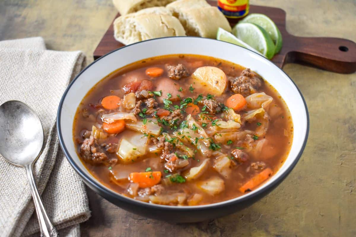 The finished soup served in a bowl with a spoon to the left side and bread, lime wedges and hot sauce in the background.