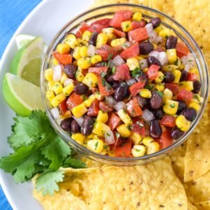 The black bean and corn salsa dip served in a glass bowl with corn tortilla chips, lime wedges and a cilantro sprig on a white plate.