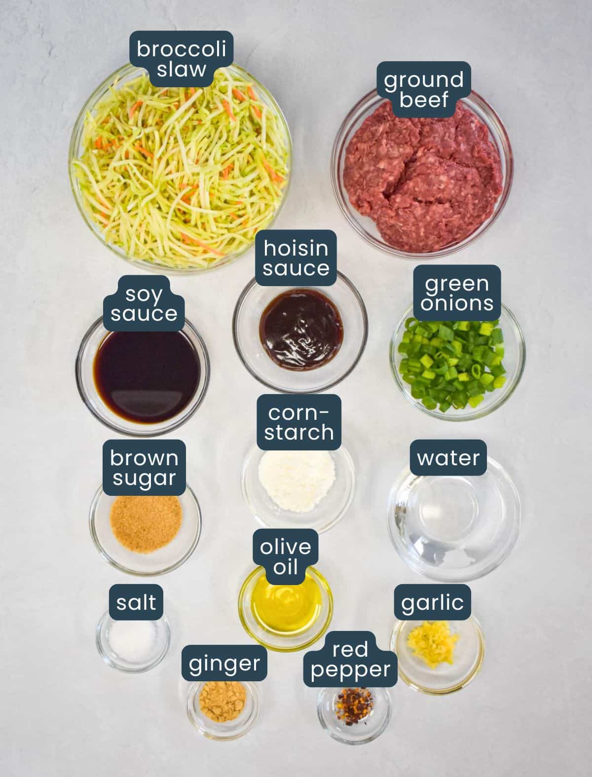 The ingredients for the dish prepped and arranged in glass bowls on a white table with each labeled with blue and white letters.