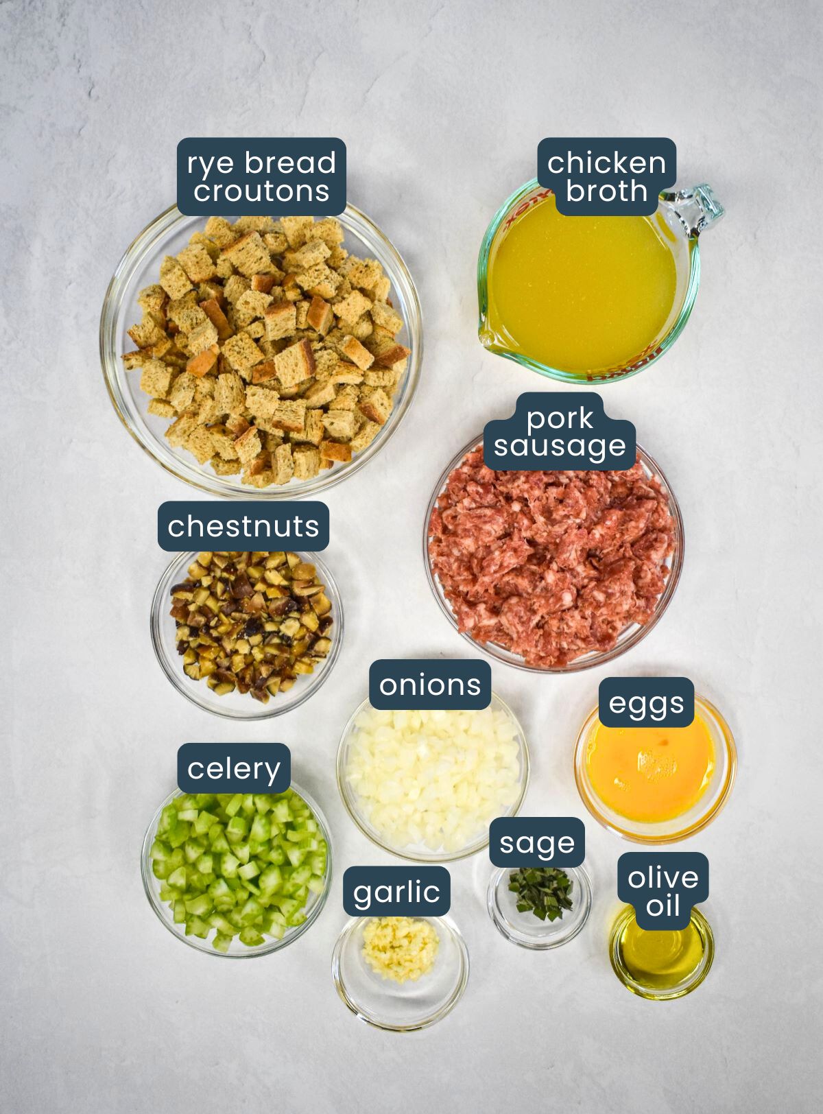 The ingredients for the stuffing prepped and arranged in glass bowls on a white table with each labeled with blue and white letters.