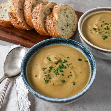 Two bowls of the creamy chestnut soup with sliced bread in the background and two spoons to the left side.