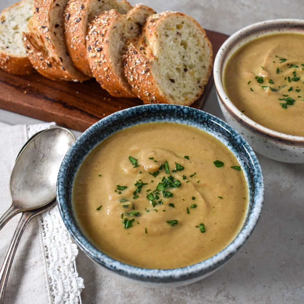 Two bowls of the creamy chestnut soup with sliced bread in the background and two spoons to the left side.