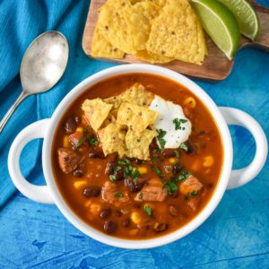 The chicken taco soup served in a white bowl garnished with parsley, sour cream, and tortilla chips with extra chips, lime wedges and a spoon in the background.