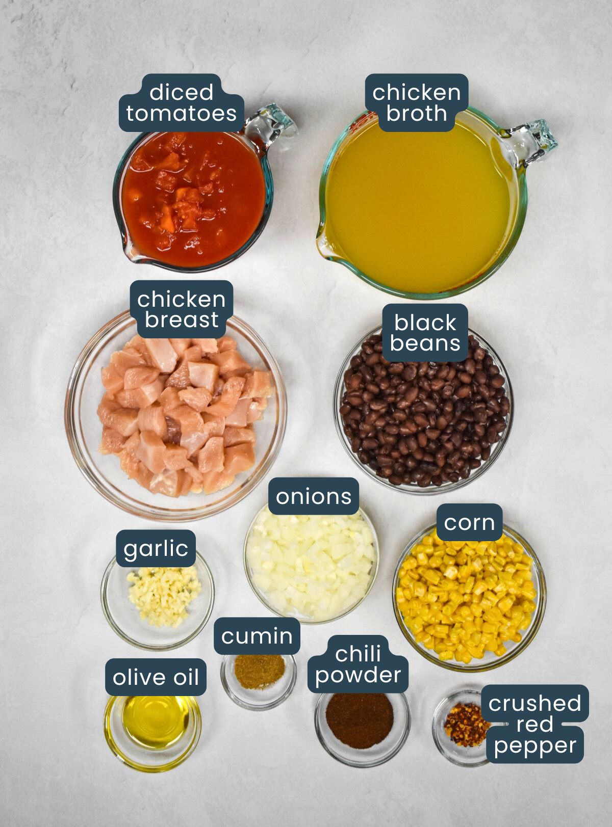 The ingredients for the soup prepped and arranged in glass bowls on a white table with each labeled with blue and white letters.