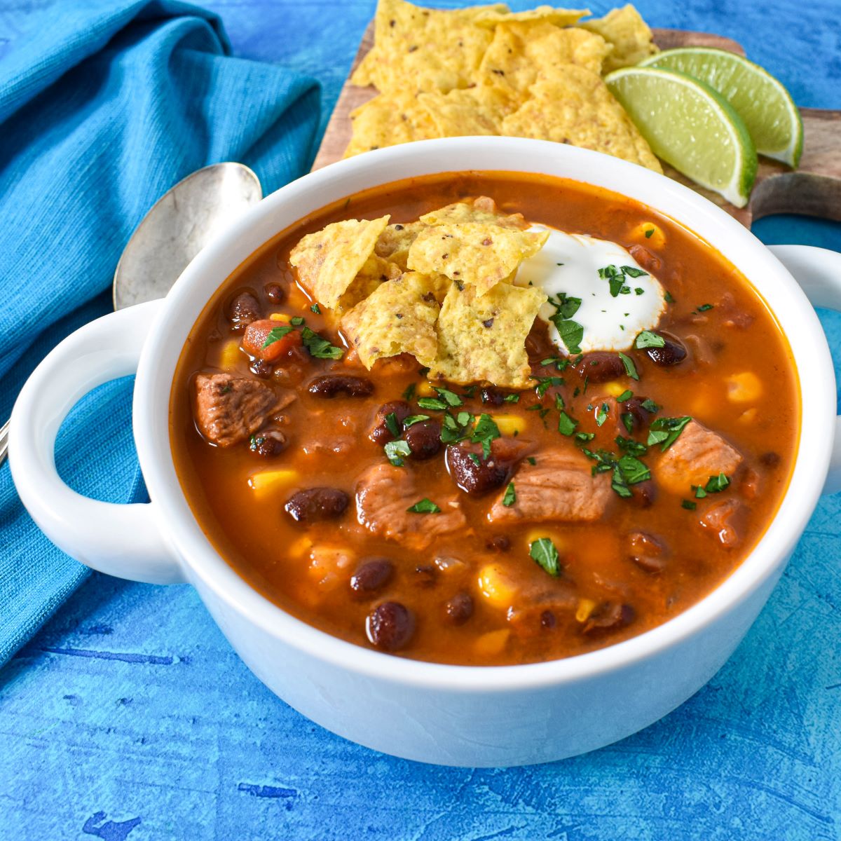 The soup served in a white bowl garnished with parsley, sour cream, and tortilla chips with extra chips, lime wedges and a spoon in the background.