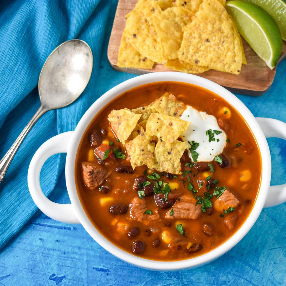 The soup served in a white bowl garnished with parsley, sour cream, and tortilla chips with extra chips, lime wedges and a spoon in the background.