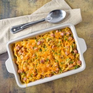 The finished baked pasta with ham in a casserole dish with a beige linen and serving spoon.