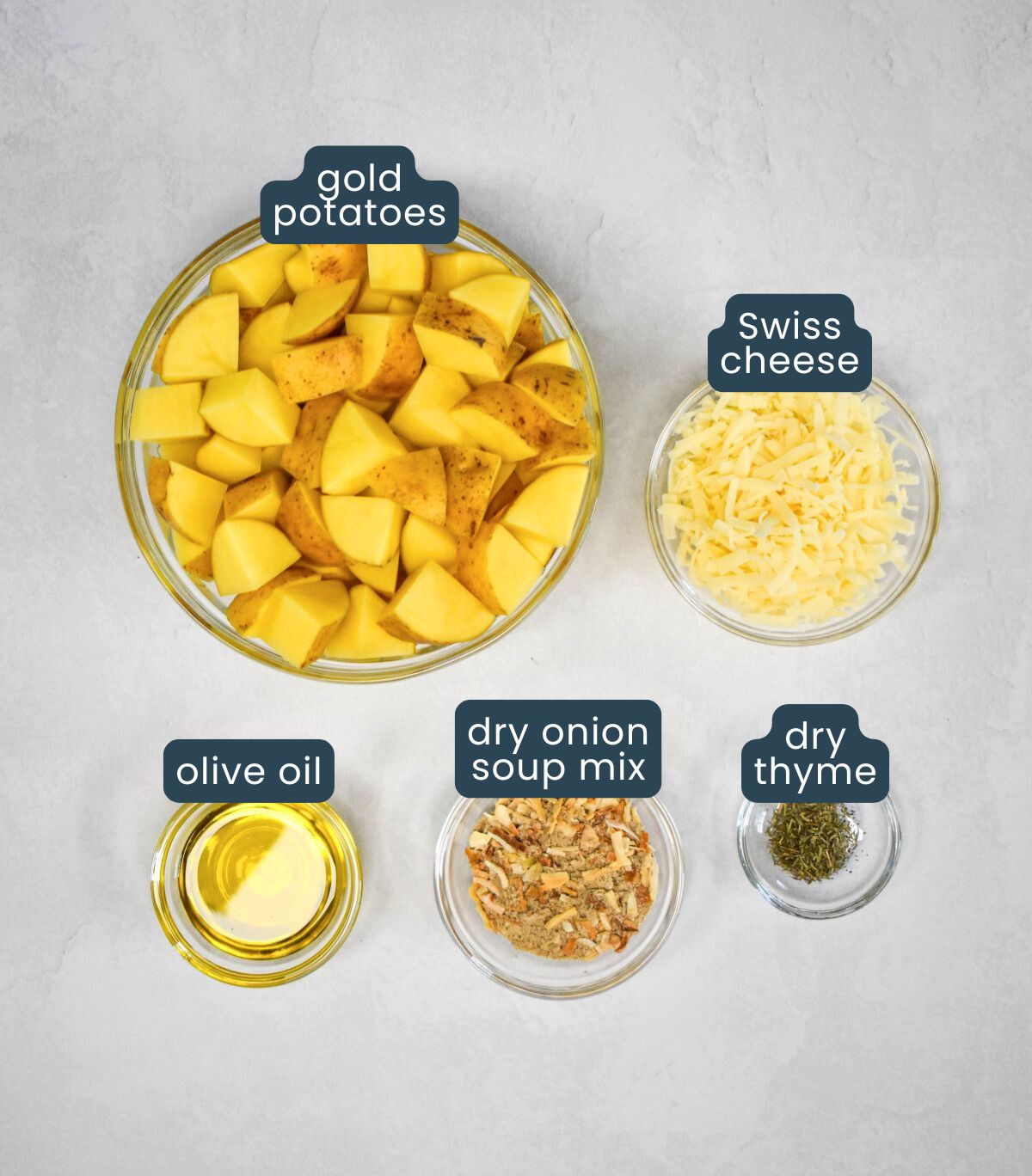 Glass bowls filled with cubed gold potatoes, shredded Swiss cheese, olive oil, dry onion soup mix, and dried thyme, arranged on a light surface.