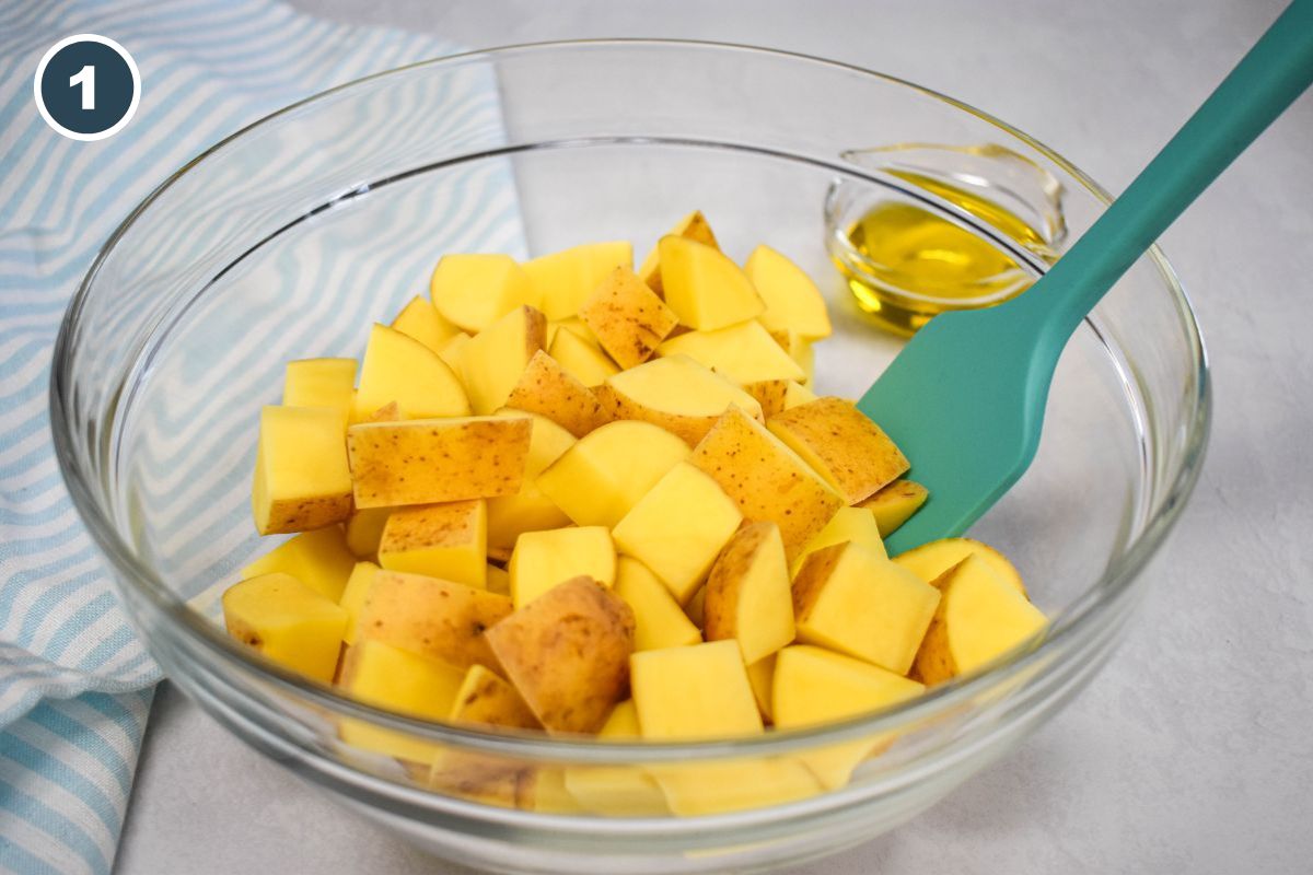 A glass bowl with cubed gold potatoes and a teal spatula, ready to be tossed with olive oil.