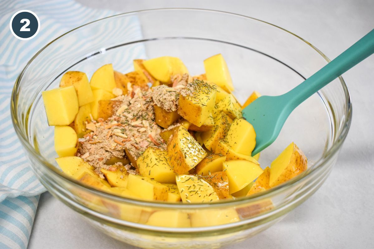 Cubed potatoes in a glass bowl, sprinkled with dry onion soup mix and dried thyme, with a green spatula.