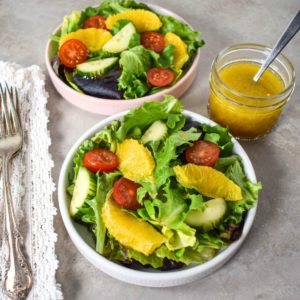 Two plates of mixed green salad with orange slices, cherry tomatoes, and a side of dressing in a glass jar.