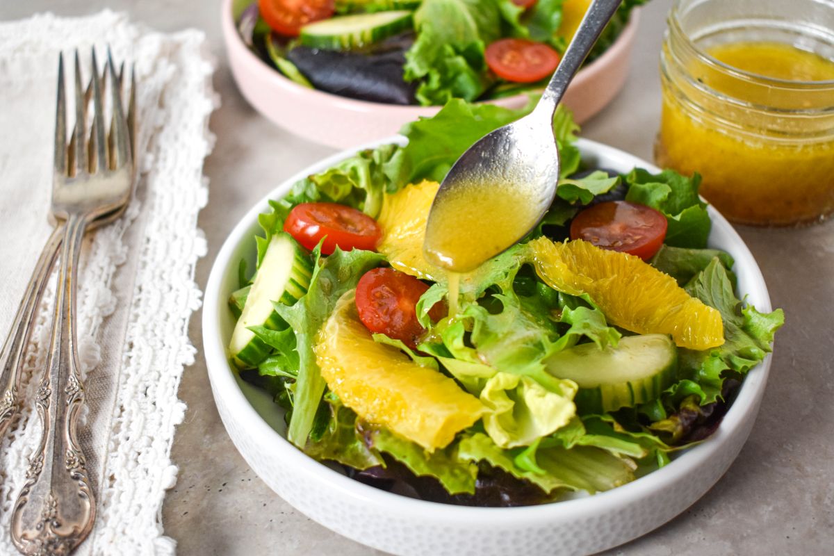 A spoon drizzling dressing over a fresh green salad with orange slices, cherry tomatoes, and cucumber.