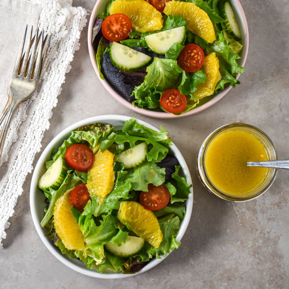 Two bowls of mixed green salad with orange slices, cherry tomatoes, and a side of dressing in a glass jar.