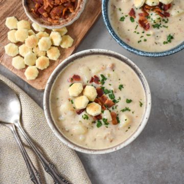 Two bowls of clam chowder garnished with crispy bacon, chopped parsley, and oyster crackers, served on a gray surface with a wooden board holding extra crackers and bacon. Two spoons rest on a beige napkin nearby.