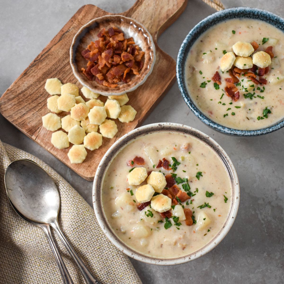 Two bowls of soup garnished with bacon, parsley, and oyster crackers, served with a wooden board holding extra crackers and bacon. Two spoons rest on a beige napkin nearby.