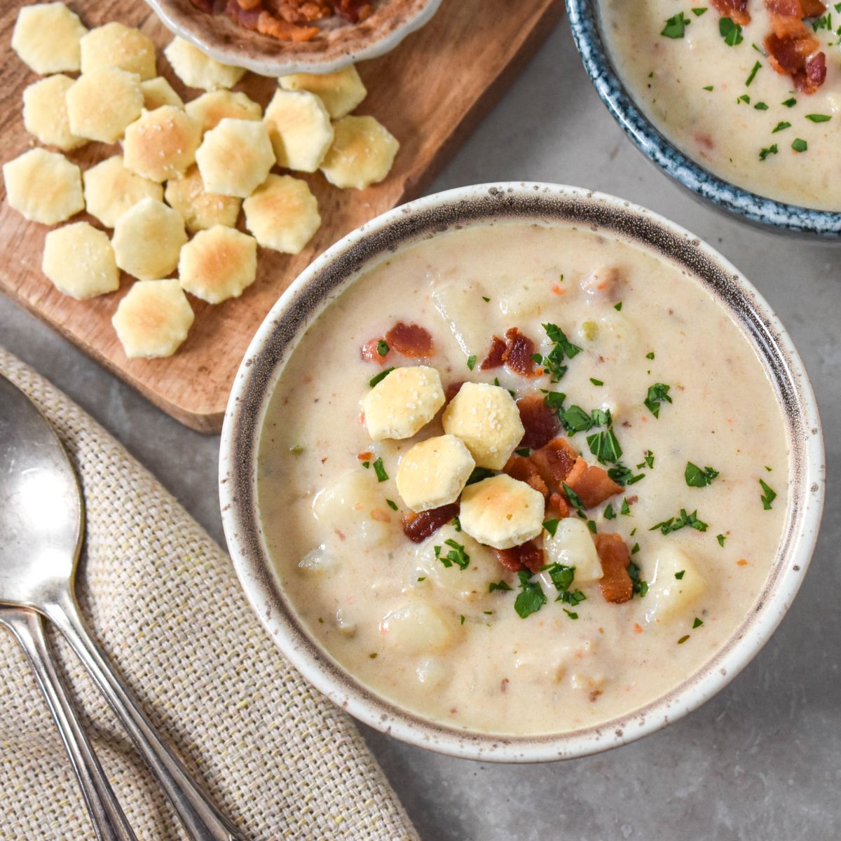 A bowl of creamy soup topped with bacon, parsley, and oyster crackers, with extra crackers and bacon on a wooden board in the background. Two spoons rest on a beige napkin nearby.