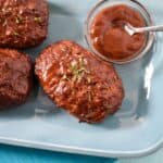 Mini turkey meatloaf with a glossy glaze, garnished with parsley, served on a blue plate with a side of sauce.