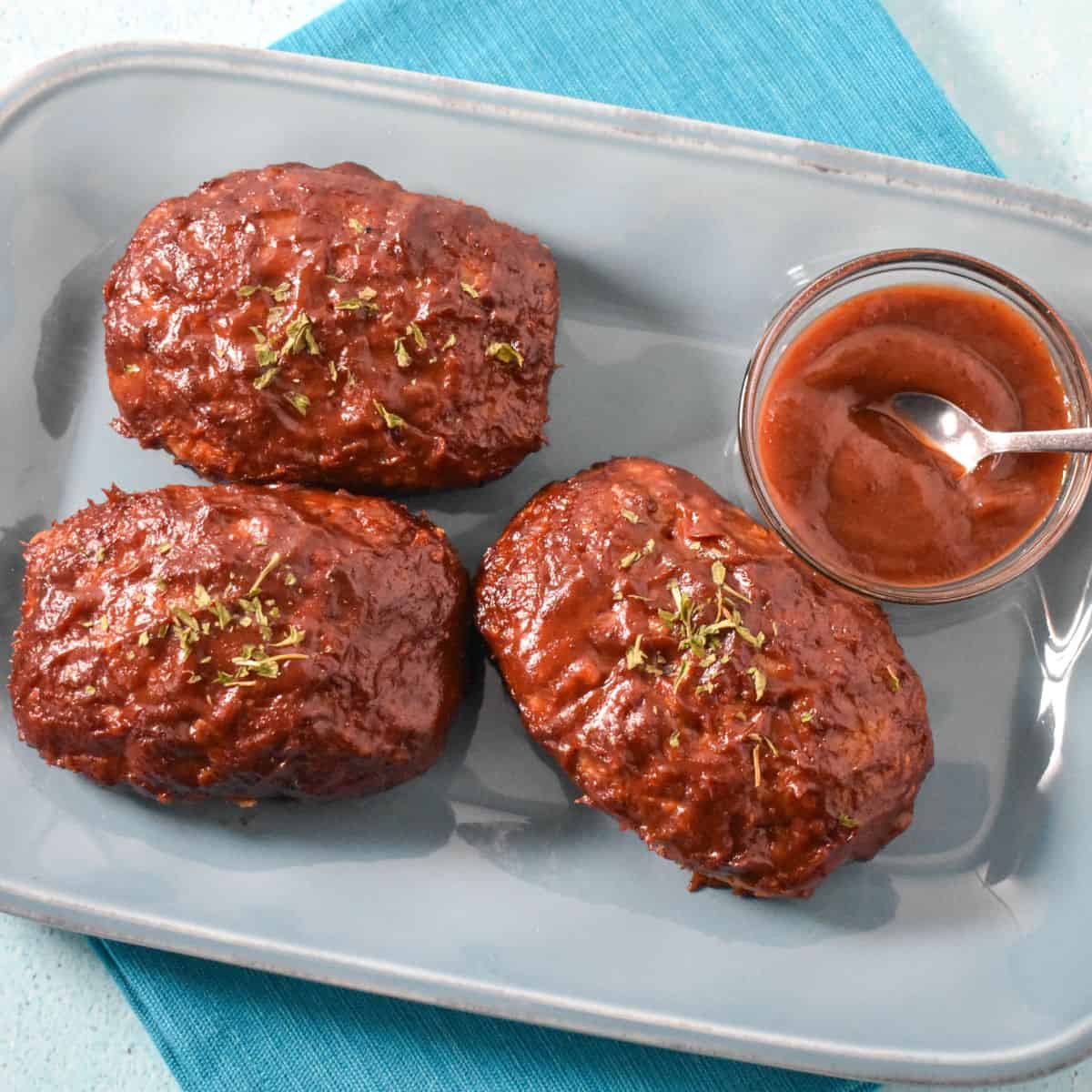 Three glazed mini turkey meatloaves garnished with parsley, served on a blue plate with a side of sauce.