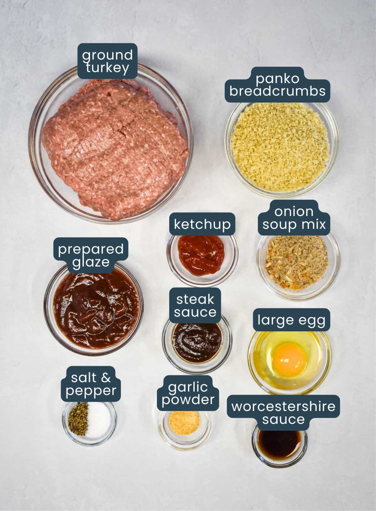 Labeled ingredients for mini turkey meatloaf, including ground turkey, panko breadcrumbs, onion soup mix, egg, ketchup, steak sauce, Worcestershire sauce, garlic powder, salt, pepper, and prepared glaze, arranged in glass bowls on a white surface.