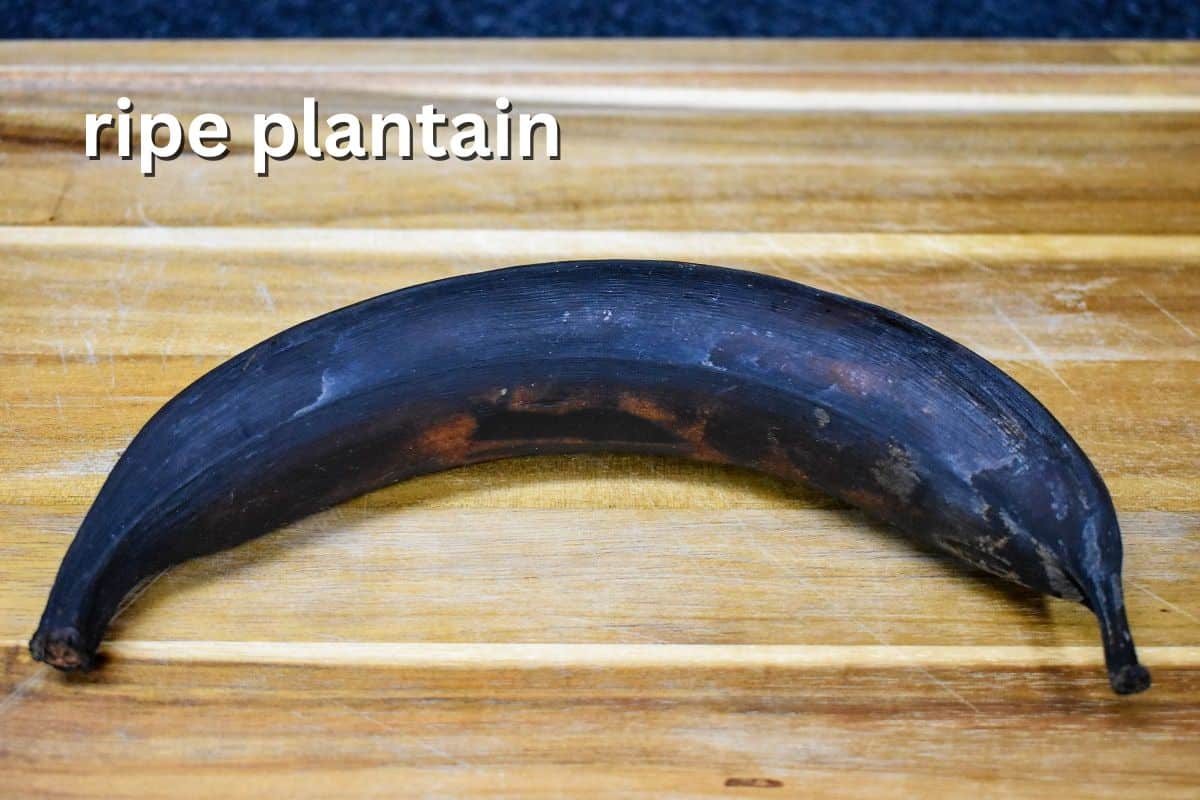 A whole black, fully ripe plantain on a wooden cutting board, labeled "ripe plantain."